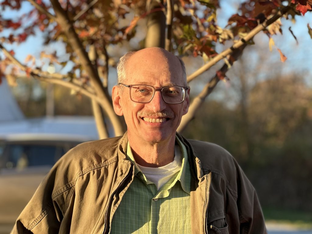 A portrait of Tom DeMatties standing outside in front of a young autumn tree and smiling at the camera.